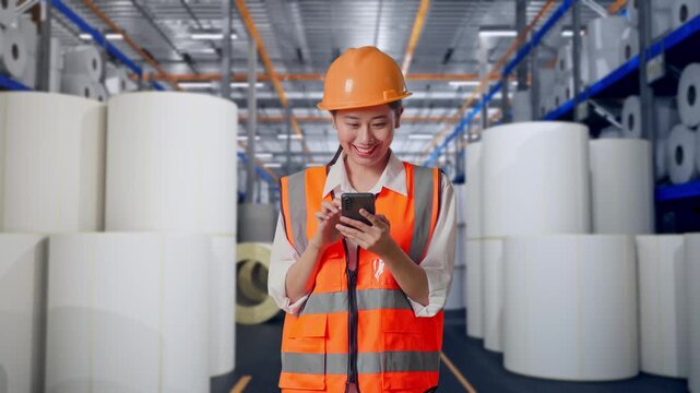 Asian Female Engineer With Safety Helmet Using Smartphone And Looking Around While Standing at Warehouse with Rows of Stacked White Polymer and Paper Rolls