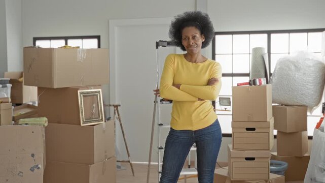 Woman standing with stacked moving boxes, arms crossed inside a building entryway, smiling; fresh start hopeful.