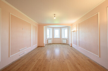 Obraz premium A spacious, empty room with light pink walls, wooden laminate flooring, a bay window with three panes, white radiators, recessed ceiling light, and wall sockets. Clean and ready for furnishing