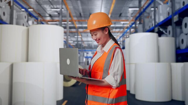 Side View Of Asian Female Engineer With Safety Helmet Working On A Laptop While Standing at Warehouse with Rows of Stacked White Polymer and Paper Rolls