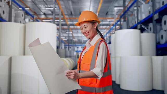 Side View Of Asian Female Engineer With Safety Helmet Looking At Blueprint In Her Hands While Standing at Warehouse with Rows of Stacked White Polymer and Paper Rolls