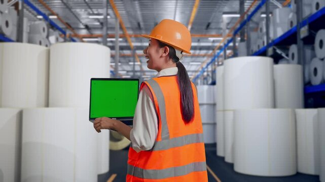 Back View Of Asian Female Engineer With Safety Helmet Working On A Green Screen Laptop And Looking Around at Warehouse with Rows of Stacked White Polymer and Paper Rolls