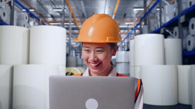 Close Up Of Asian Female Engineer With Safety Helmet Working On A Laptop While Standing at Warehouse with Rows of Stacked White Polymer and Paper Rolls