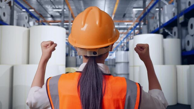 Close Up Back View Of A Female Engineer With Safety Helmet Raising Her Hands Celebrating While Working at Warehouse with Rows of Stacked White Polymer and Paper Rolls