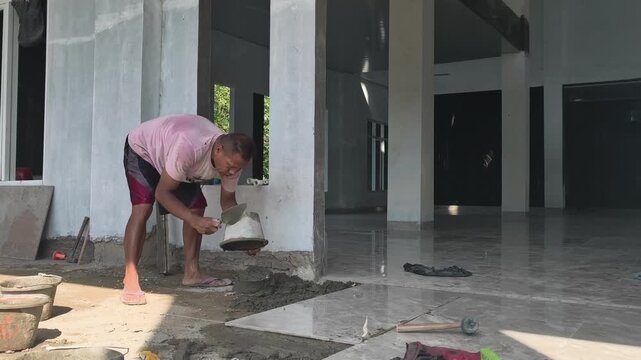 Construction worker pouring wet cement mixture on a floor surface in preparation for floor tile installation. Base layer preparation work.