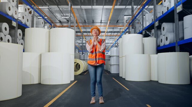 Full Body Of Asian Female Engineer With Safety Helmet Pray For Something While Standing at Warehouse with Rows of Stacked White Polymer and Paper Rolls