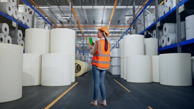 Full Body Back View Of Asian Female Engineer With Safety Helmet Working On A Green Screen Smartphone And Looking Around at Warehouse with Rows of Stacked White Polymer and Paper Rolls