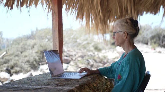Mature woman sits at a table under a palapa working on a computer at a beach in Baja California, Mexico surrounded by nature.