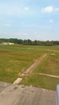 Airplane lands on the runway at Milan Malpensa Airport in Italy. The view from the window reveals green fields of grass and airport buildings under a blue sky dotted with white clouds.