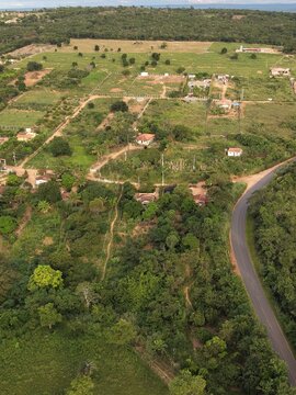 Captura a&eacute;rea mostrando uma paisagem rural tranquila com pequenas propriedades, casas isoladas, vegeta&ccedil;&atilde;o nativa densa e uma estrada sinuosa sob a luz do dia.
