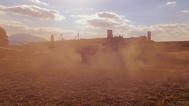 Heavy agricultural machinery tilling the soil, kicking up dust in the golden evening light