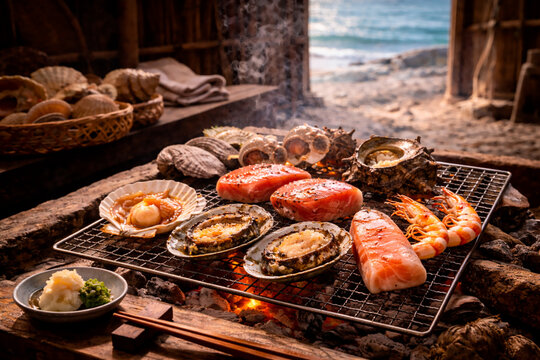 A variety of fresh seafood, including scallops, abalone, prawns, and salmon fillets, grilling over a charcoal fire at a rustic seaside Ama hut (Ama-goya).