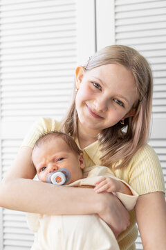 Portrait of a smiling blonde preteen sister gently holding her newborn brother with a pacifier in his mouth against a light background