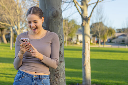 teen girl looking at her phone while leaning against a tree in a suburban park