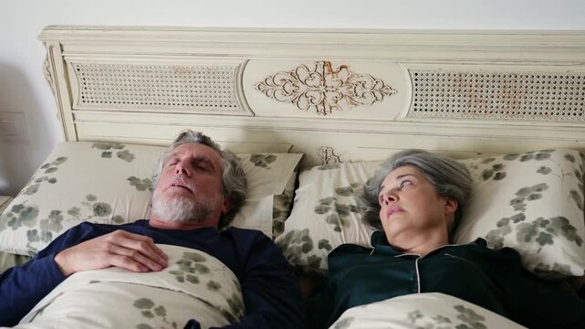 Annoyed senior woman covering her ears with a pillow because of her husband snoring loud at night