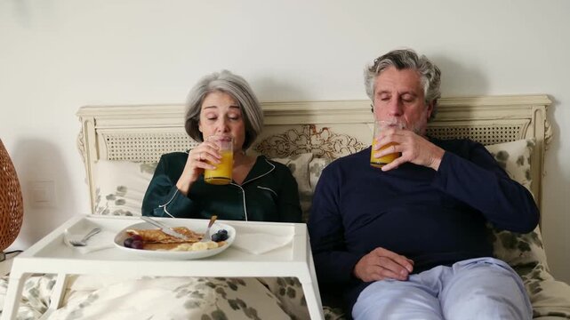 Happy senior couple in pajamas talking and drinking orange juice while eating breakfast in bed