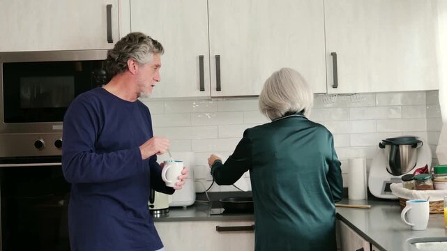 Lovely elderly couple in pajamas making breakfast and talking together in their cozy kitchen