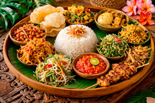 A vibrant and traditional Nasi Campur platter featuring a central mound of steamed white rice surrounded by various Indonesian side dishes, including chicken satay, sambal, spicy green beans, shredded