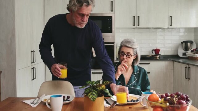 Happy senior man and woman in pajamas enjoying breakfast and talking together in the morning at home