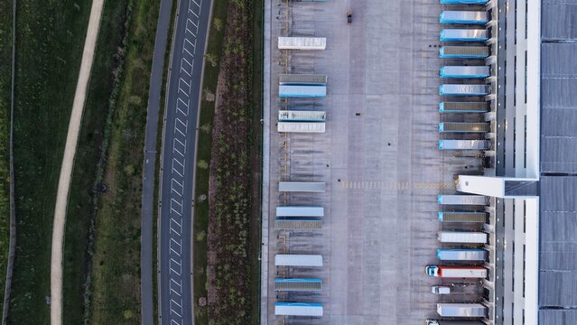 Birmingham, United Kingdom - 21 April 2026: Aerial view of the Amazon EMA4 UXD1 fulfillment center with rows of blue semi-trailers parked at loading docks and a nearby road.