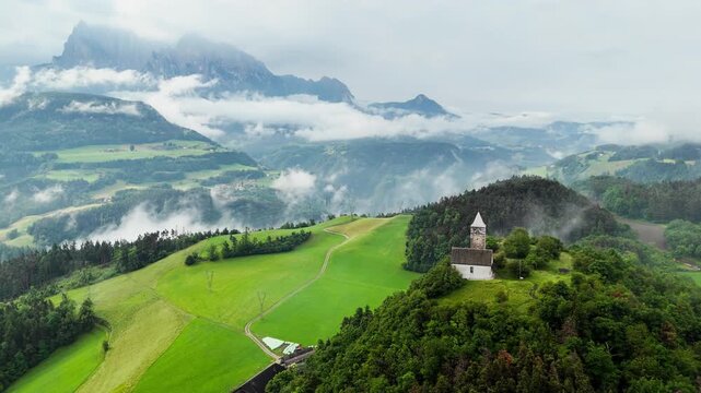 Aerial view of chapel on hilltop in South Tyrol, Dolomites, Italy. Alpine landscape with green fields, forest and low clouds drifting over mountains