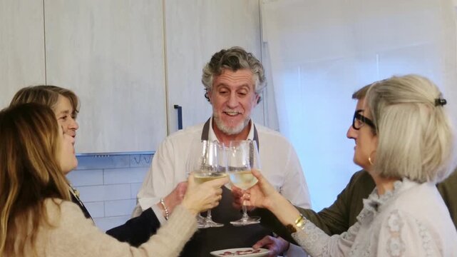 Cheerful group of mature friends enjoying a house party, clinking glasses with white wine