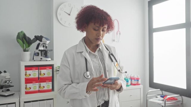 Woman doctor with stethoscope tapping smartphone in clinic building exam room, microscope and medical supplies visible, clock on wall; calm focus.