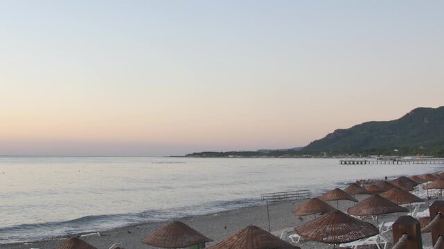 Tranquil sunrise over a pebble beach with rows of straw umbrellas and a calm sea in the background.
