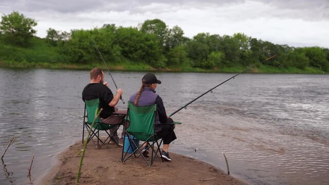 Calm man and woman sitting on folding chairs on a riverbank, patiently holding fishing rods and waiting for a bite, enjoying a quiet outdoor hobby together on a cloudy day