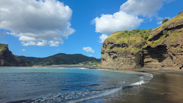 15 Oct 2025, New Zealand, A scenic view showcases a black sand beach meeting the turquoise ocean under a partly cloudy sky.