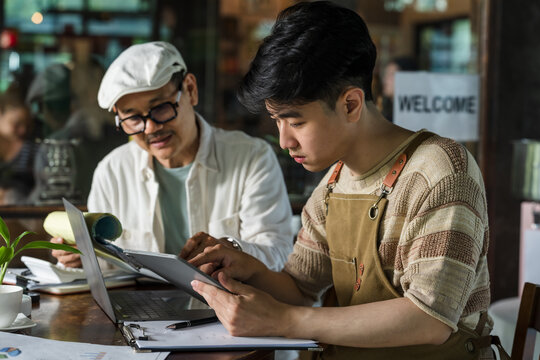 Asian father guides teenage son using tablet and laptop for cafe task review, document study, order planning, and family restaurant management lesson during real business training session today.
