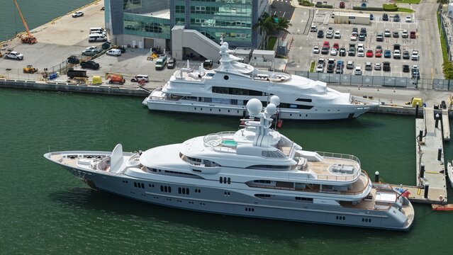 Riviera Beach, United States - 15 April 2026: Aerial view of luxury superyachts docked at the marina with a modern building and parking lot in the background.