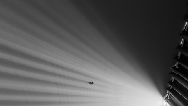 Abstract view of black and white ceiling showing radiating light streaks from window blinds, with a small smoke detector centered in frame