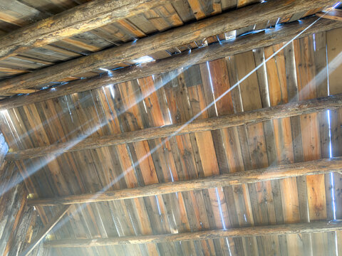 Looking up at a rustic barn ceiling with weathered log rafters, wooden planks, and dramatic light rays piercing through gaps