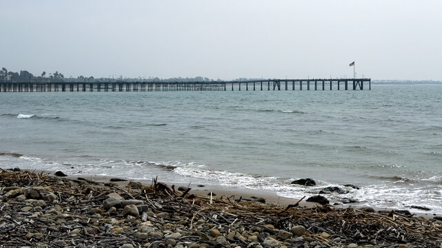 Rocky debris-strewn shoreline with driftwood, pebbles, and gentle waves, Ventura Pier silhouetted in distant hazy background