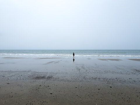 Anonymous lone figure, standing silhouetted at water's edge on vast flat wet beach, tiny against expansive grey sky and calm ocean