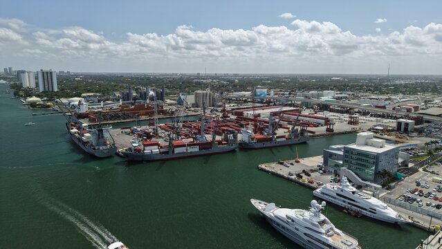 Riviera Beach, United States - 15 April 2026: Aerial view of the industrial port area with cargo ships, shipping containers, and luxury yachts docked under a partly cloudy sky.