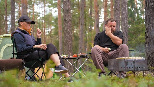 Relaxed man and woman sitting on camping chairs, eating snacks and waiting for the barbecue to cook while enjoying their vacation in a beautiful pine forest with a tent in the background