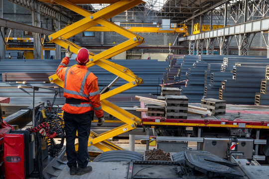 Worker operating yellow scissor lift beside transport trailer and stacked steel beams inside industrial warehouse with overhead cranes and machinery