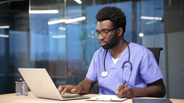 Portrait of confident African American doctor in blue coat works on computer in office sitting at desk at workplace in hospital clinic. Medical worker physician busy with paperwork, looking at camera