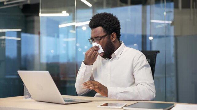 Allergic african american businessman sneezes and wipes his nose with a handkerchief while sitting at desk at workplace in modern business office. Unwell male worker suffering from seasonal allergies
