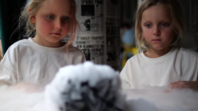 Two fascinated young girls with blonde hair watching a smoking cauldron with wonder. Thick white smoke billows from the bowl, creating a mysterious and magical atmosphere during the science show