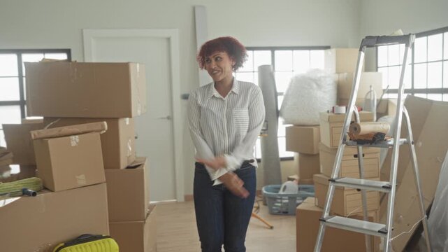 Woman in striped shirt spreads arms among stacked cardboard boxes, ladder and yellow suitcase inside building, smiling and looking up with open hands; moving day joy.