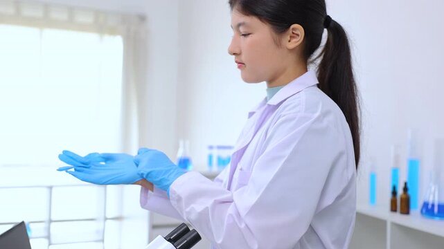 Teen girl in white coat wearing gloves before research task in a modern lab. Science, laboratory, scientist, experiment, and research express lab readiness and scientific process, curiosity.