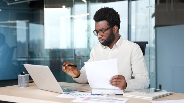 Joyful african american businessman is satisfied with the results of a financial report looking at documents and a computer screen sitting in business office. Manager is happy with positive indicators