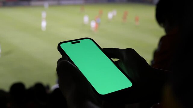 Silhouette hands holding a smartphone with a green screen mockup at a stadium, blurred live soccer match in the background, sports betting and live streaming concept