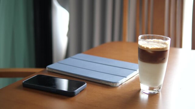 Cinematic close-up of layered iced coffee latte on a wooden table with smartphone and tablet, modern remote work and lifestyle concept