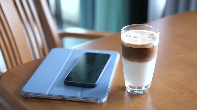 Cinematic shot of iced coffee latte with layers on a wooden table next to a tablet and smartphone, modern remote work and lifestyle concept
