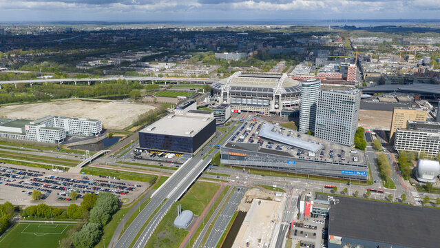 Amsterdam, Netherlands - 19 April 2026: Aerial view of Johan Cruijff Arena surrounded by modern office buildings, parking lots, and urban infrastructure under a partly cloudy sky.