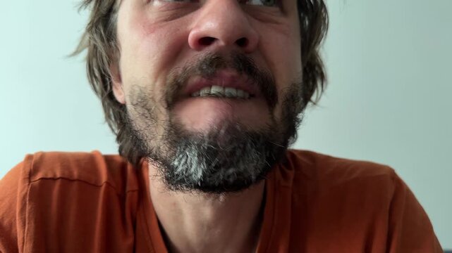 A bearded man nervously bites his lower lip in a close-up shot. Subtle facial tension and downward gaze suggest anxiety, discomfort, or inner hesitation indoors